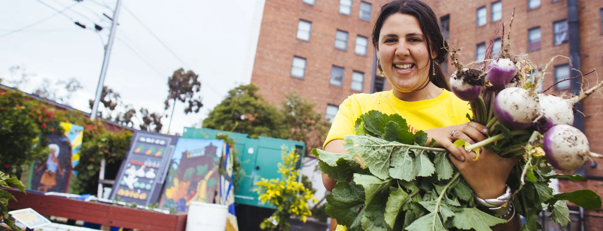 A farmer presents a crop from an urban plot in front of a brick building