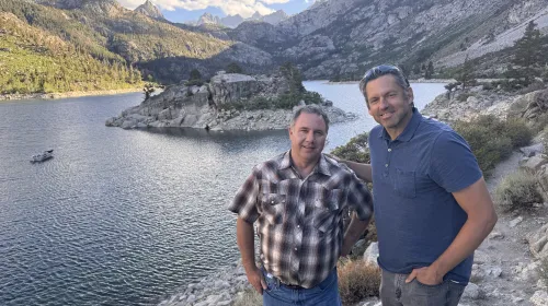 Two men stand at overlook above Sierra lake with peaks in the distance