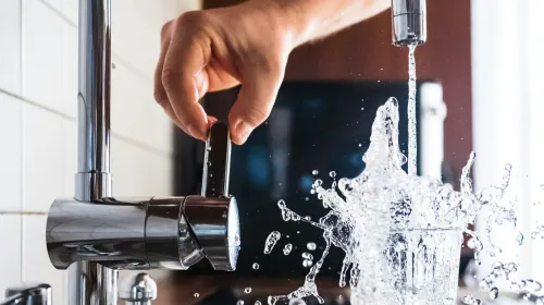 Hand turns on a tap with water splashing out of a glass
